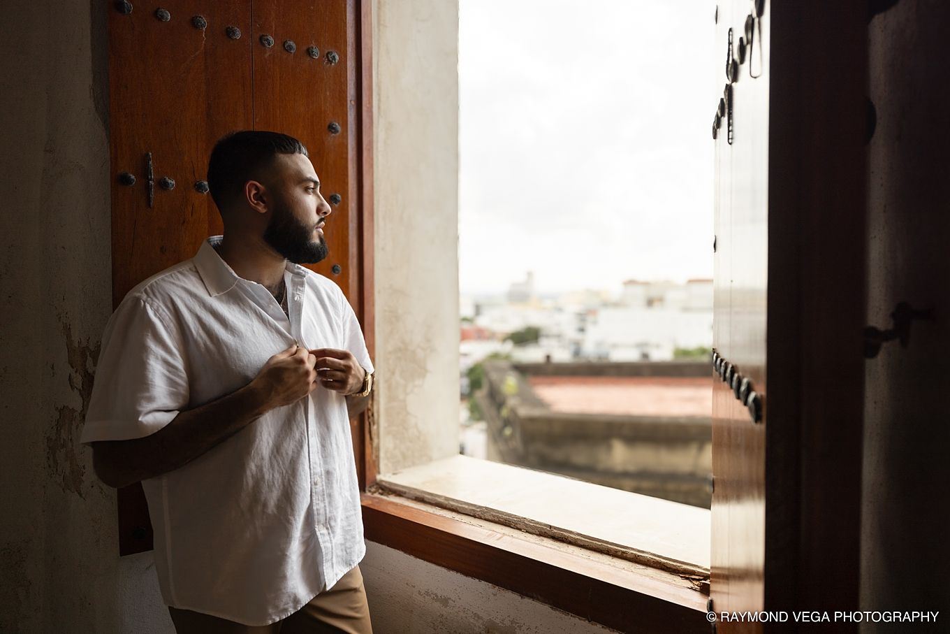 A bearded man in a white shirt gazes out an open window from a rustic balcony with a city view.