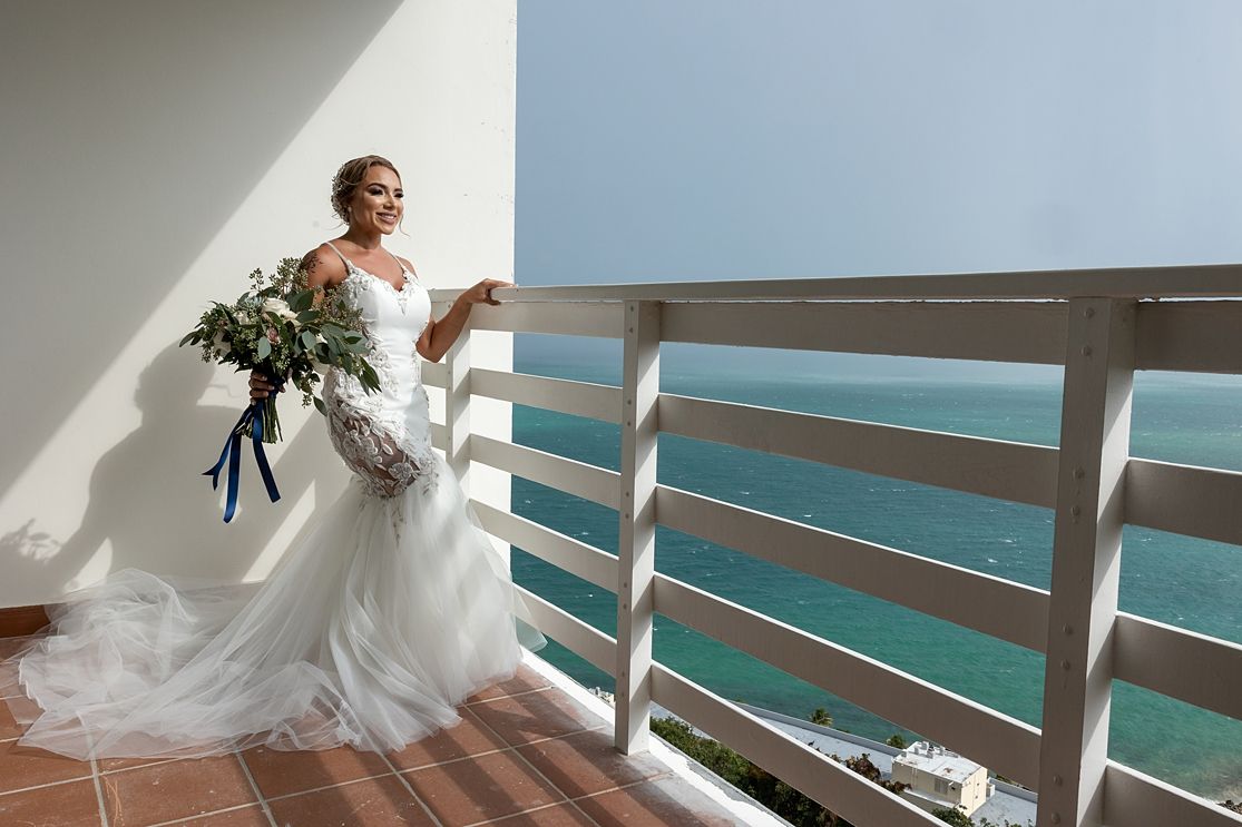Bride in white lace gown holding bouquet on oceanview balcony with turquoise water backdrop on wedding day. Hotel El Conquistador