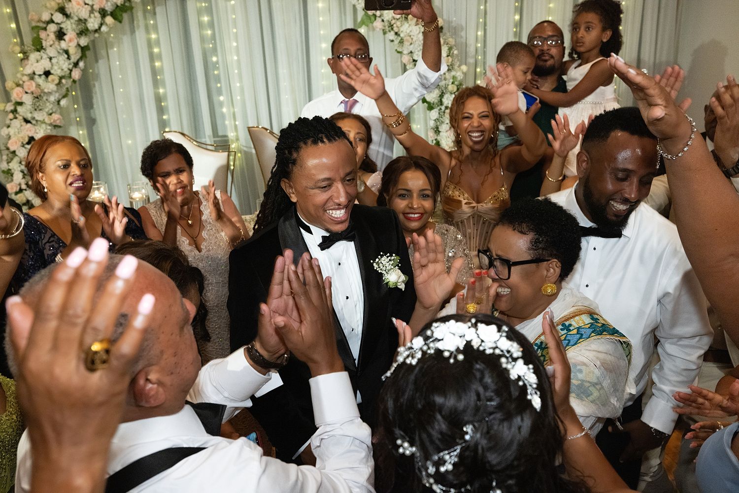 Joyful wedding celebration with guests cheering and clapping around a smiling couple in elegant attire at a decorated reception.