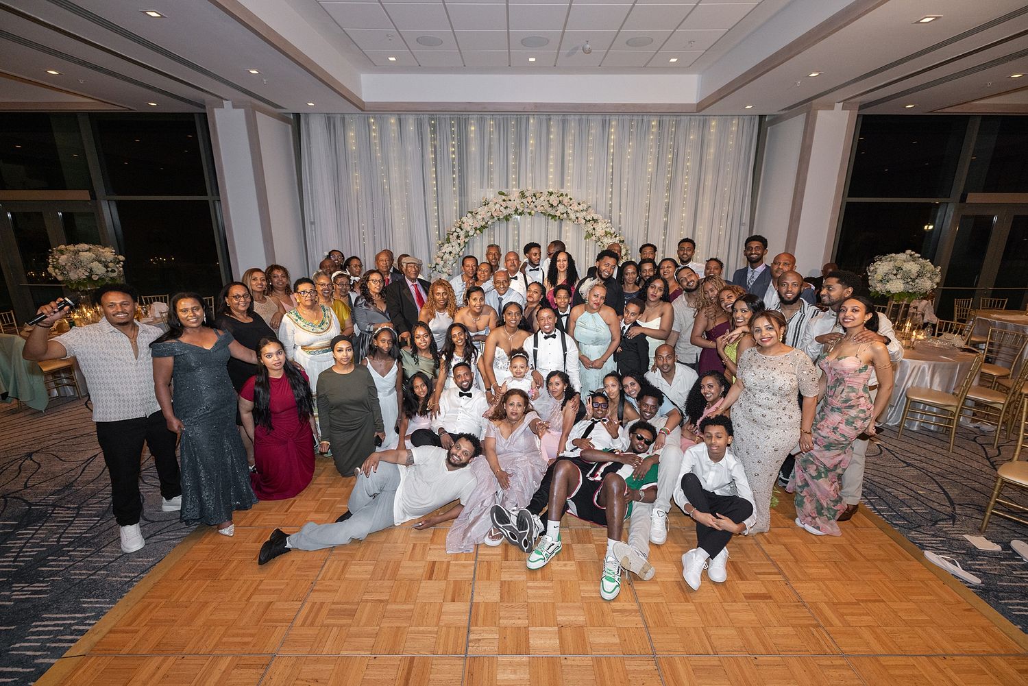 Large group photo of wedding guests posing together in an elegant ballroom with floral arch backdrop.