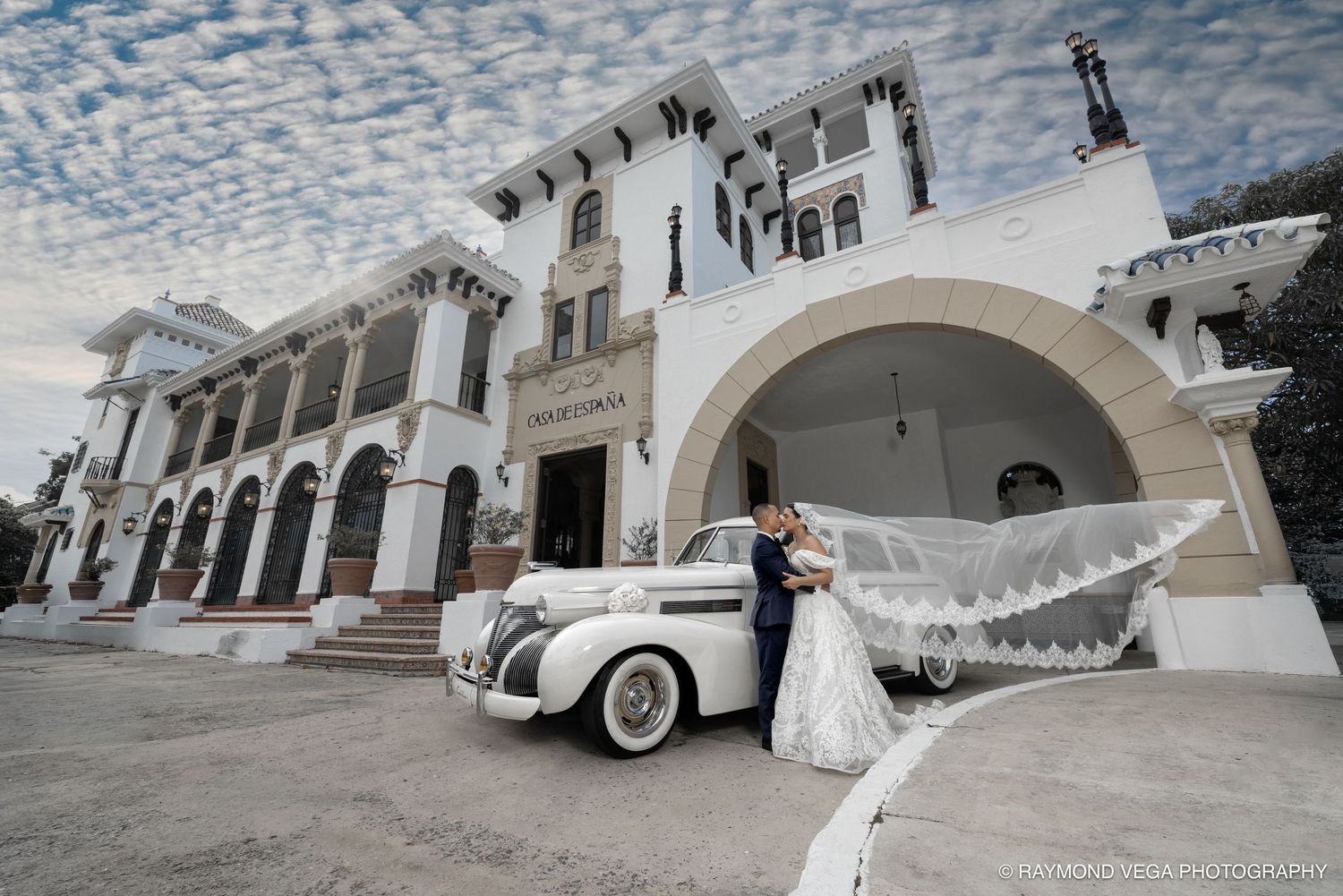 Bride and groom posing with a vintage white car in front of an elegant white building on their wedding day.