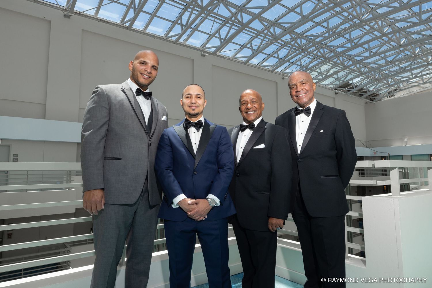 Four men in formal tuxedos and suits pose together smiling inside a modern venue with a glass ceiling.