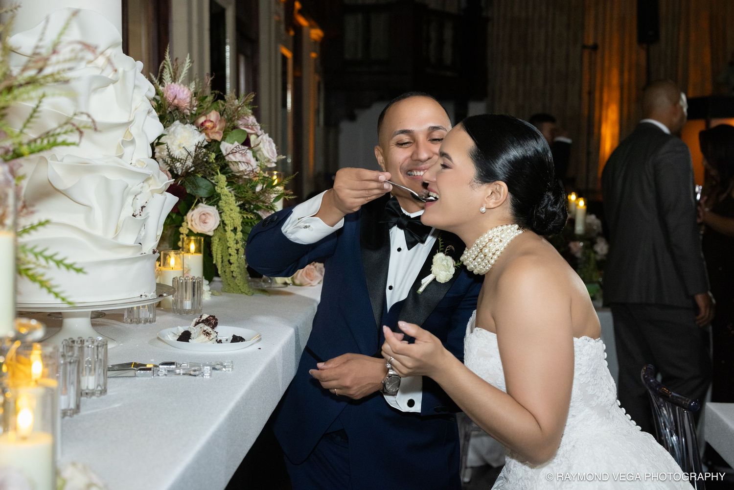 Bride and groom sharing wedding cake at reception, groom feeding bride in elegant venue with white floral arrangements.