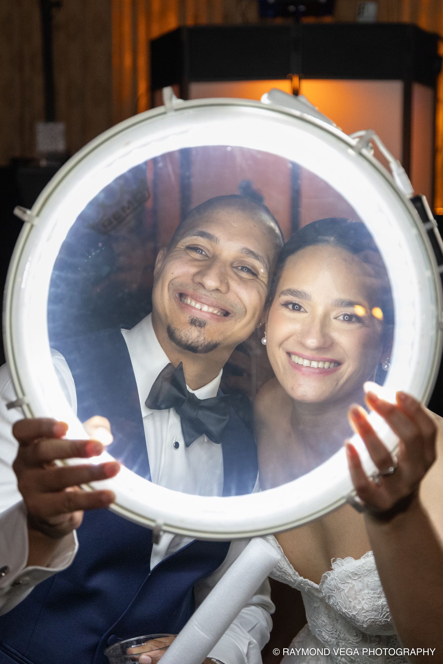 Bride and groom smiling through illuminated drum hoop at wedding reception, captured by Raymond Vega Photography.