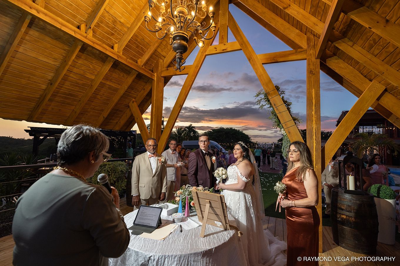 Outdoor wedding ceremony under wooden pavilion at sunset with couple exchanging vows and guests watching.