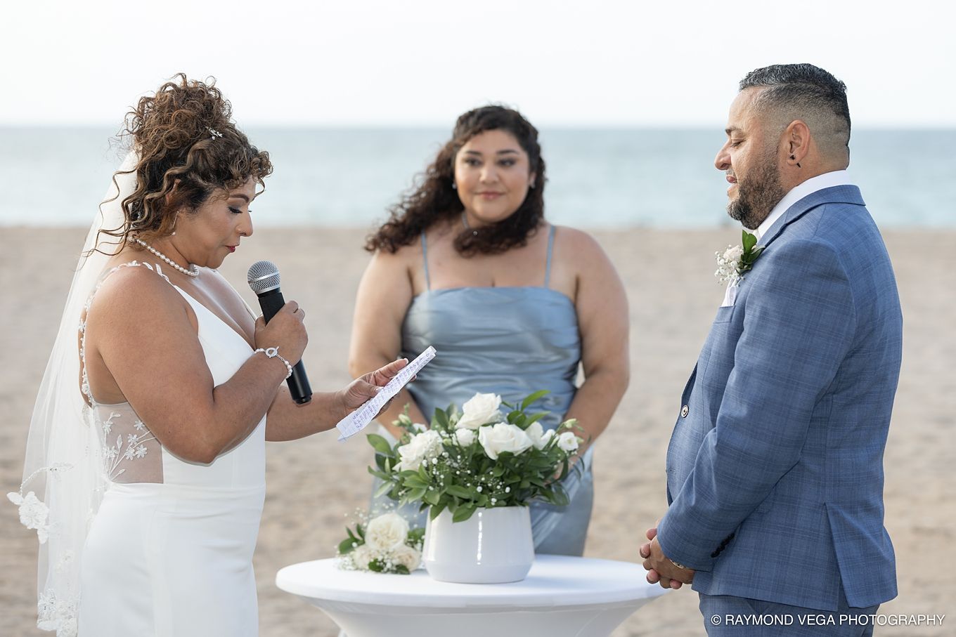 Bride reads vows to groom at romantic beach wedding ceremony with bridesmaid officiating, ocean backdrop.