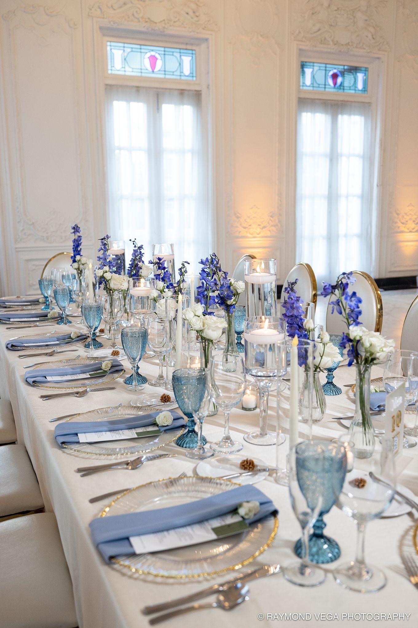 Elegant wedding reception table with blue floral centerpieces, blue glassware, and silver chargers in a white ornate ballroom. Liliana Albiz