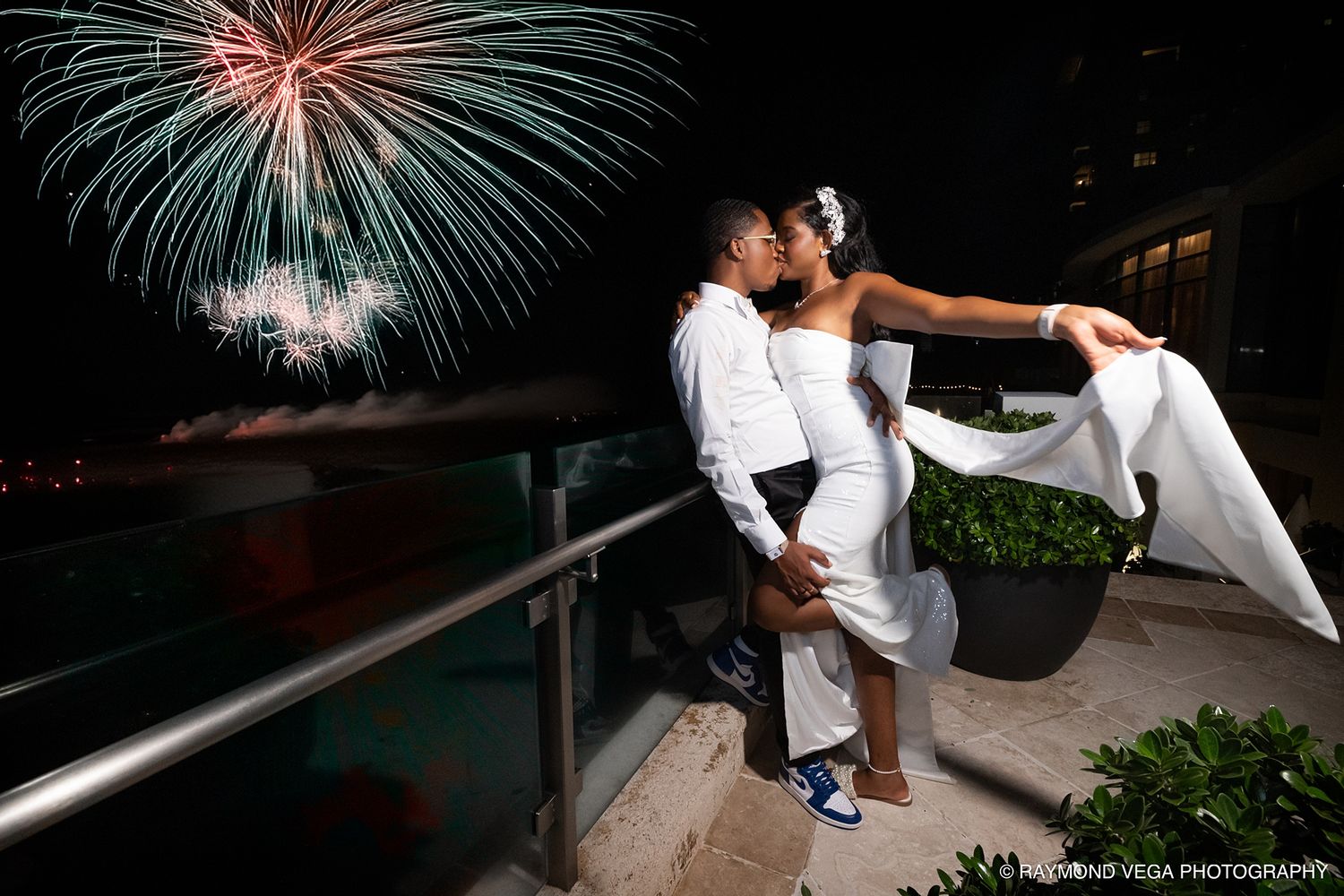 Newlywed couple sharing a romantic kiss on a balcony with colorful fireworks lighting the night sky behind them.