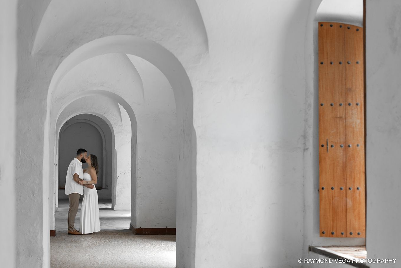 Couple sharing a romantic kiss framed by white arched hallway in elegant Mediterranean-style venue.