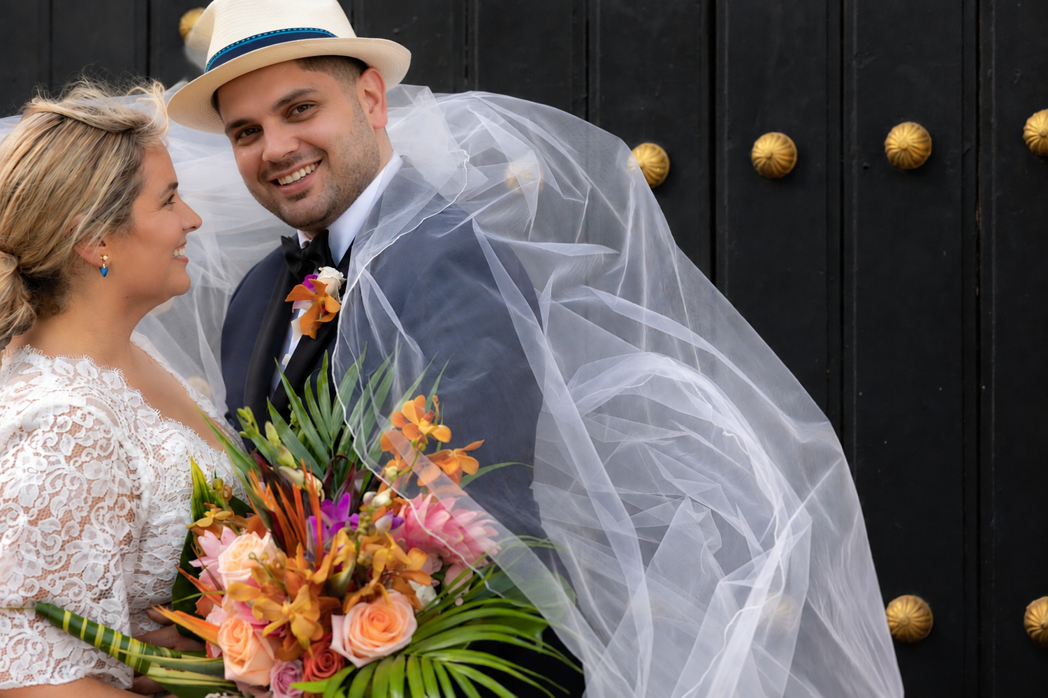 Bride and groom smiling together, veil flowing, holding vibrant tropical bouquet before ornate dark doors.
