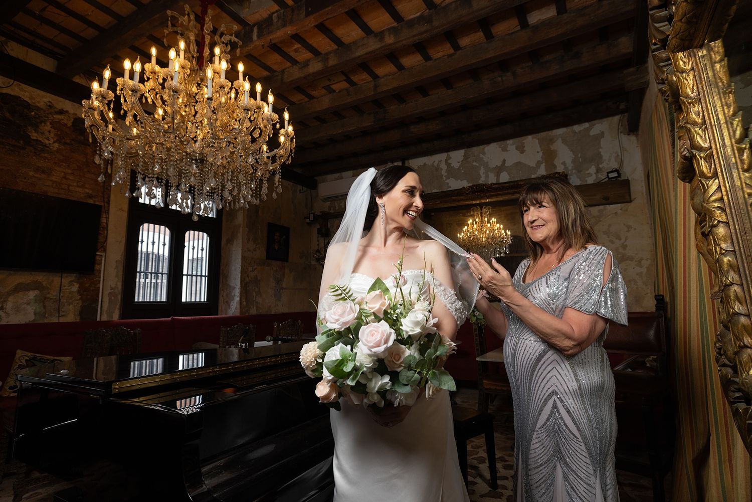 Bride in white gown holds floral bouquet as woman adjusts veil in ornate chandelier-lit wedding venue.