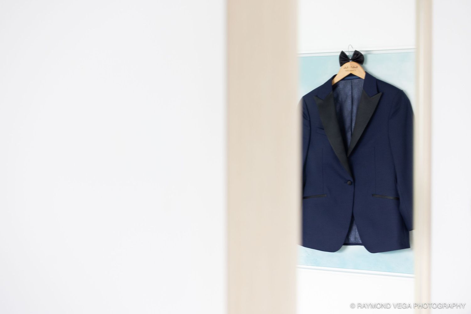 Navy blue tuxedo jacket hanging on a wooden hanger in a bright white doorway, perfect for a formal wedding.