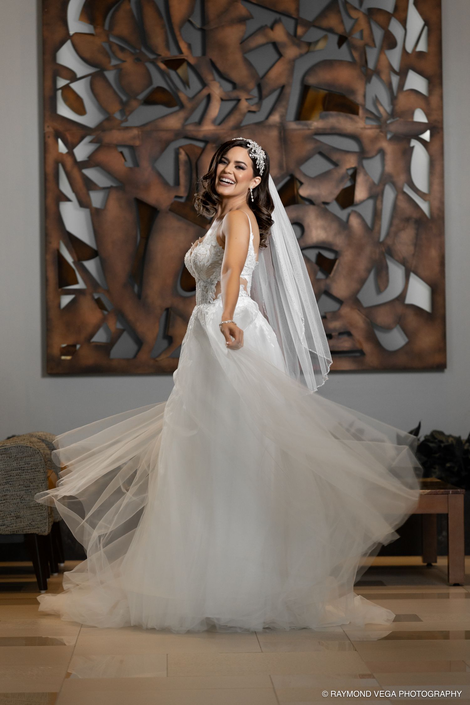 Radiant bride in a flowing white tulle ball gown and veil, laughing joyfully before an ornate bronze wall art.