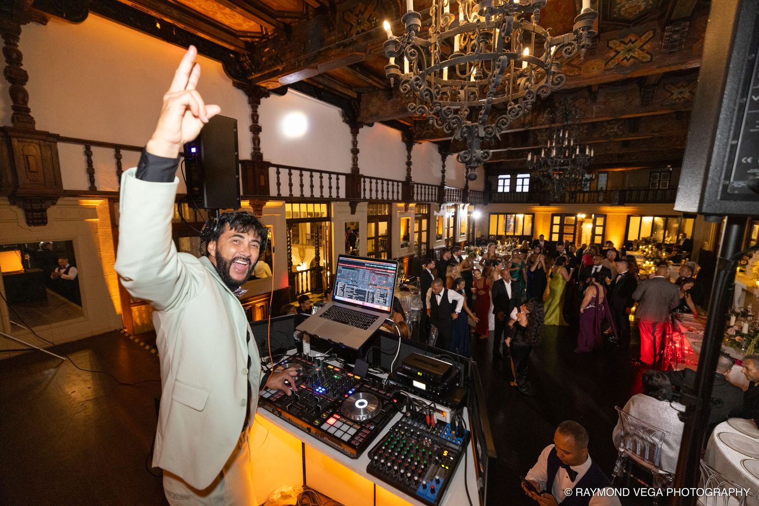 DJ celebrates at turntables during lively wedding reception in elegant venue with chandelier and dancing guests.