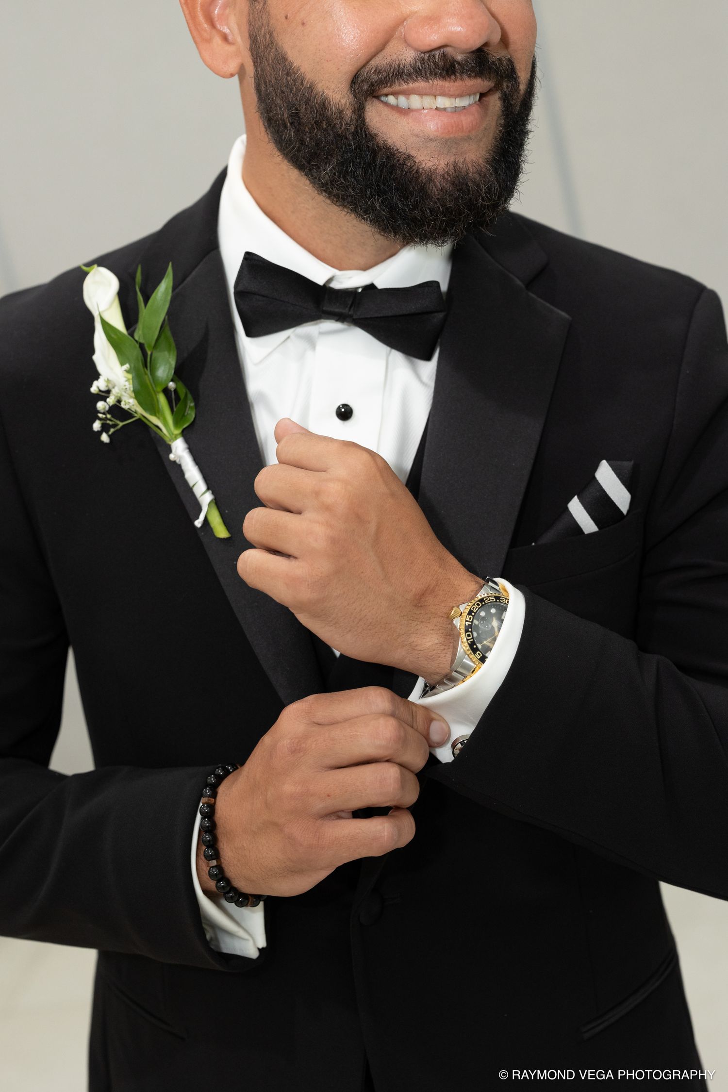 Smiling groom in black tuxedo with bow tie, gold Rolex watch, and white calla lily boutonniere.