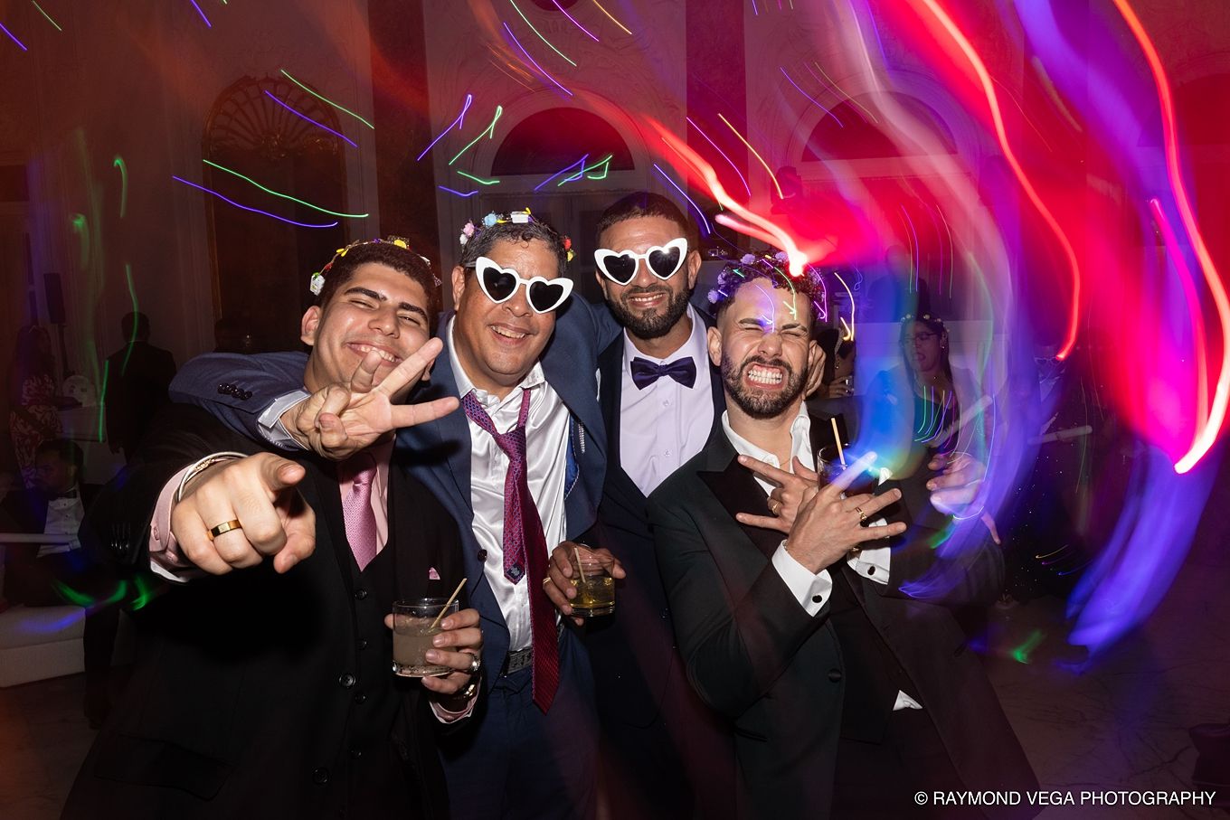 Four men in suits wearing fun heart-shaped sunglasses celebrate at a wedding reception with colorful lights. Antiguo Casino Puerto Rico