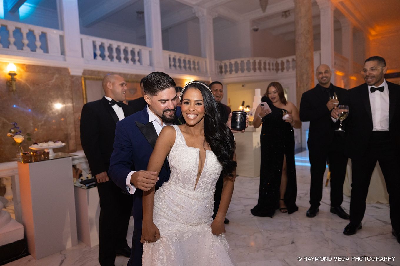 Bride in lace gown and groom share first dance at elegant wedding reception with guests celebrating in background.