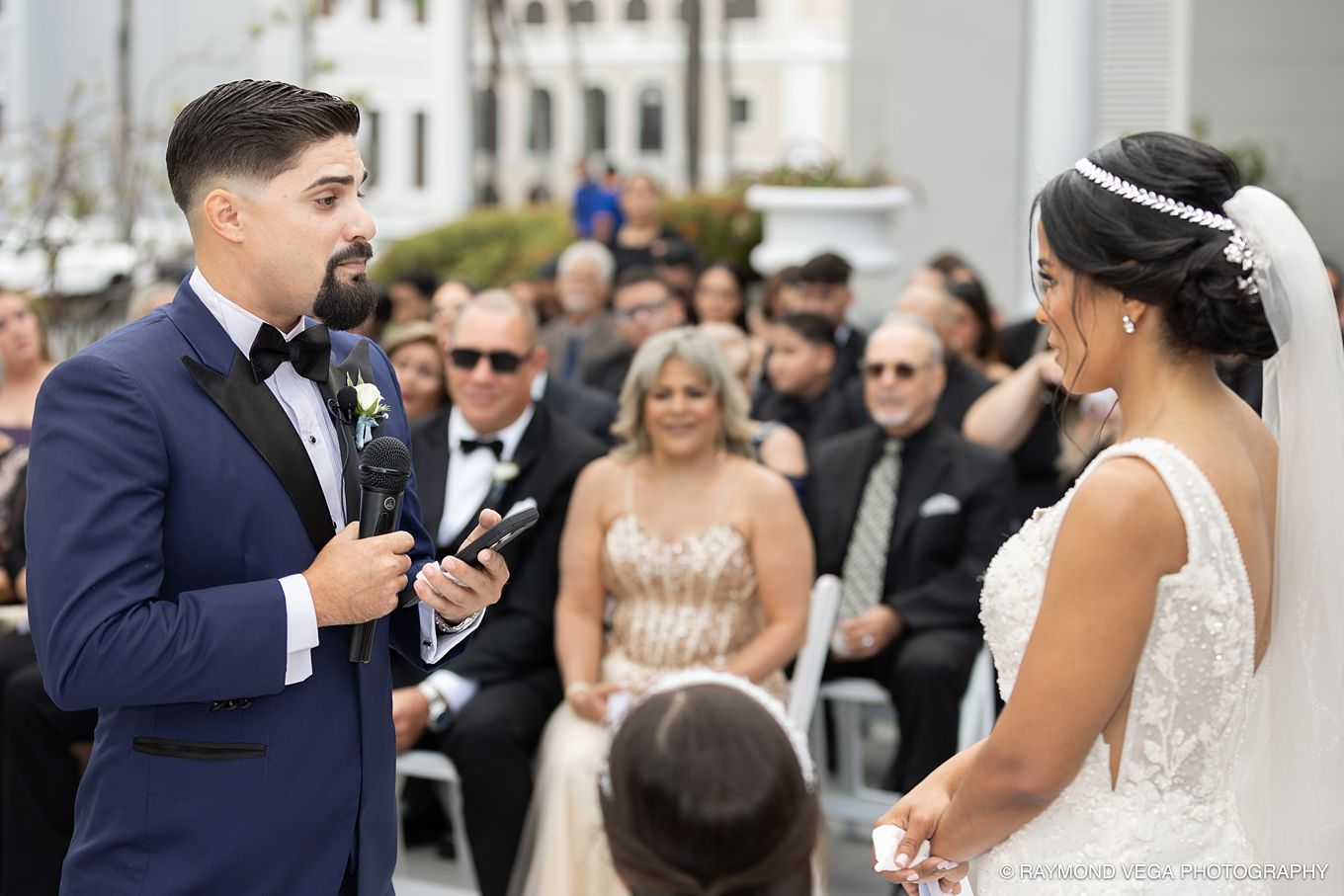 Groom in navy suit reads vows to bride in white gown during outdoor wedding ceremony with guests seated behind. Antiguo Casino Puerto Rico