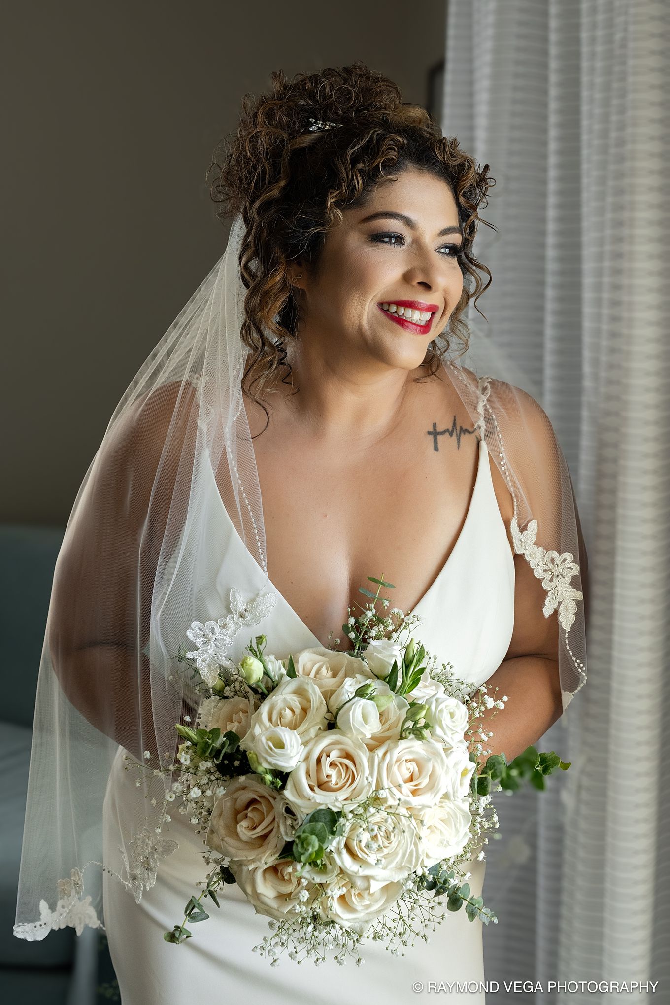 Bride in white gown holds ivory rose bouquet, wearing veil and red lipstick, smiling near window on wedding day.