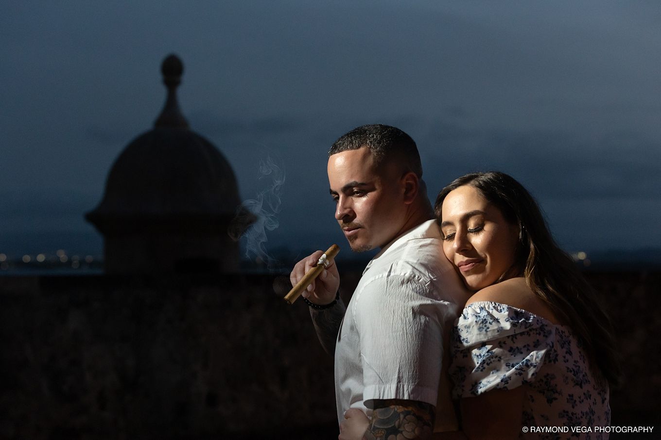 Couple poses romantically at dusk near historic dome, man smoking cigar, woman leaning close, city lights in background. - Love Story