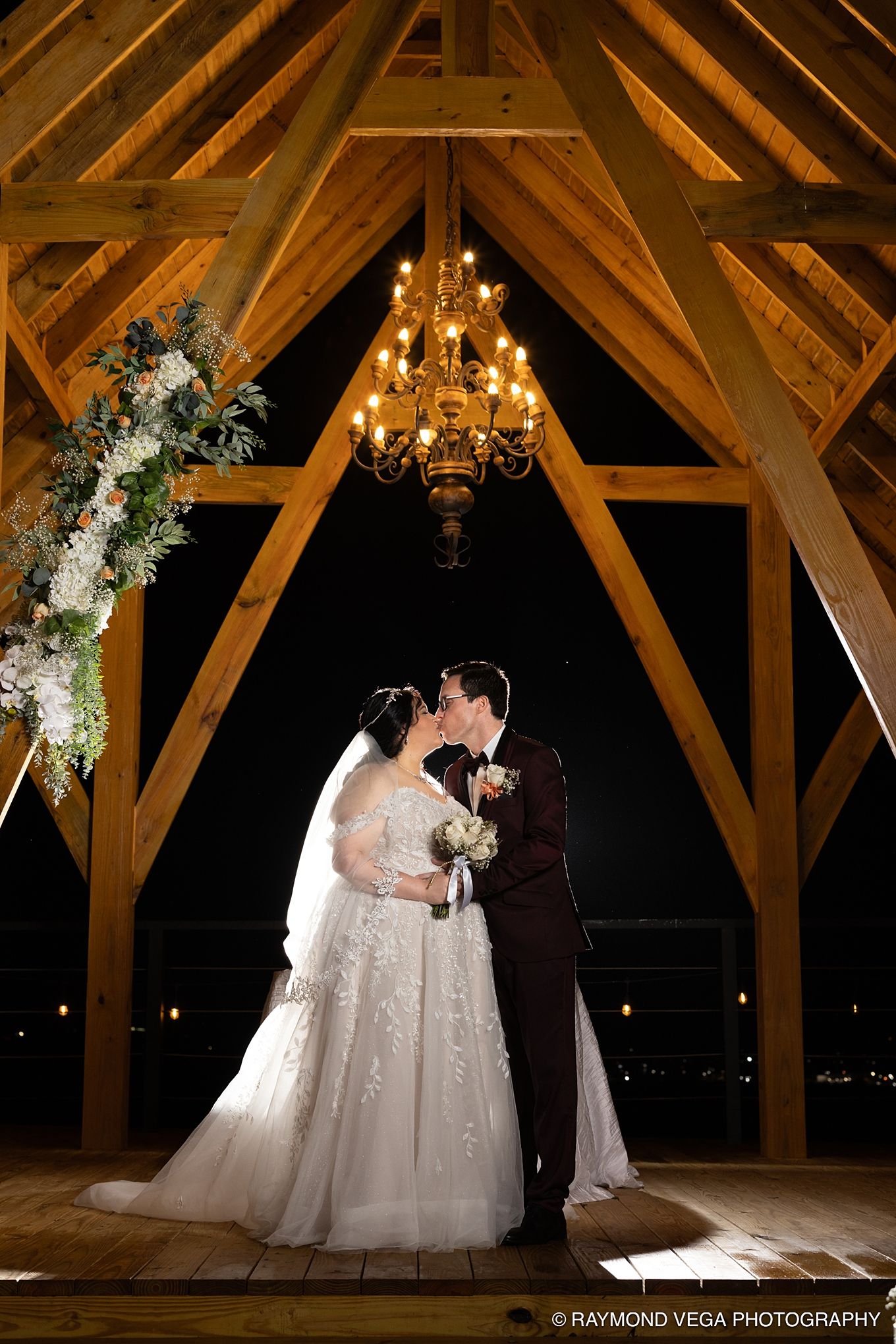 Bride and groom share a kiss under a chandelier in a wooden barn venue at night, with floral arrangements nearby.