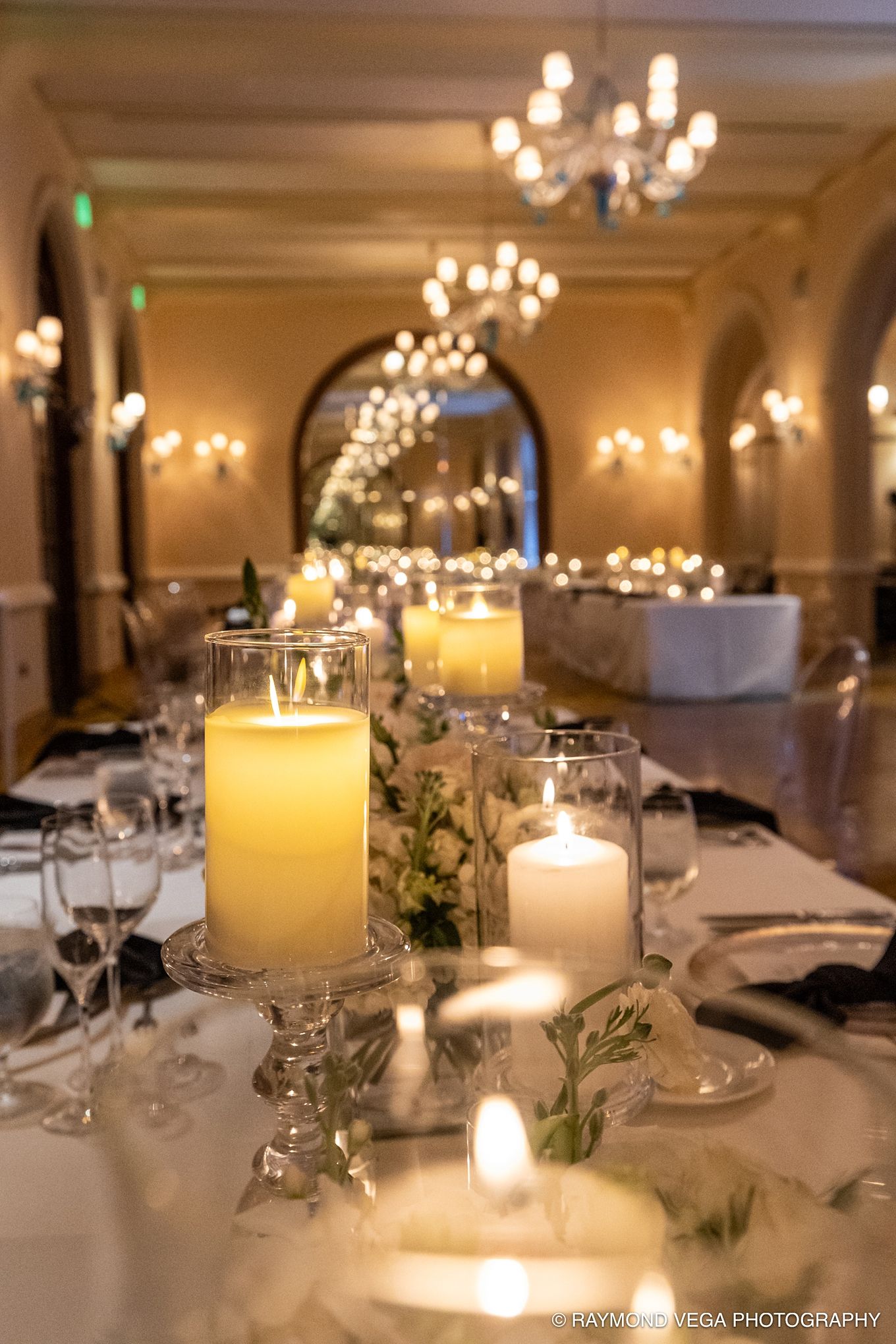 Elegant wedding reception table with glowing pillar candles in glass holders, floral greenery, and warm chandelier lighting.