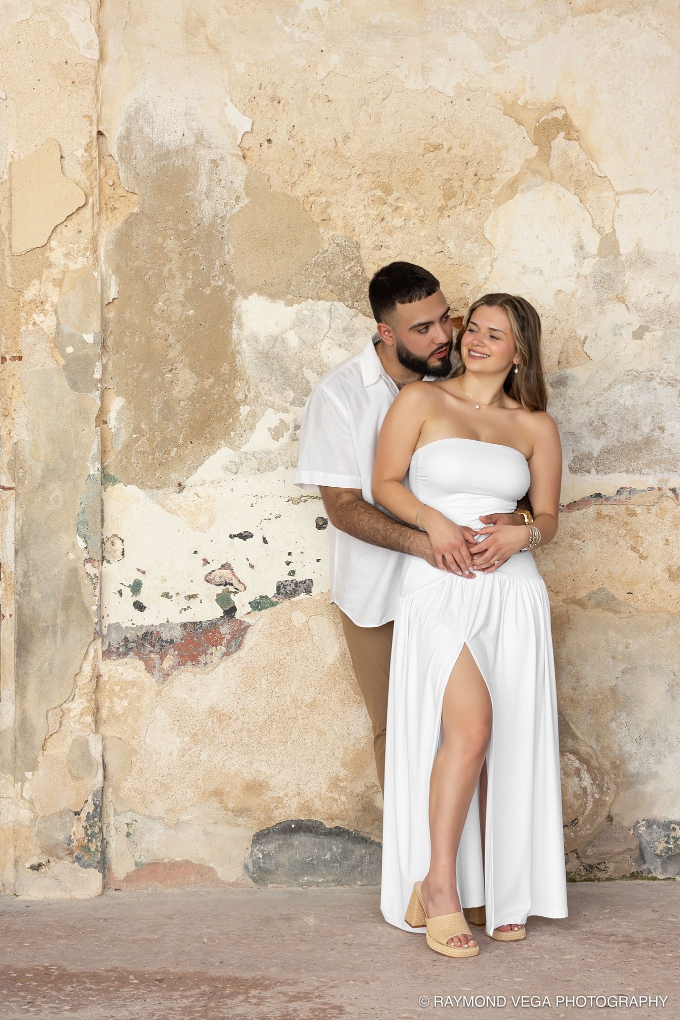 Couple in white outfits embracing against a rustic textured wall in a romantic engagement photo session.