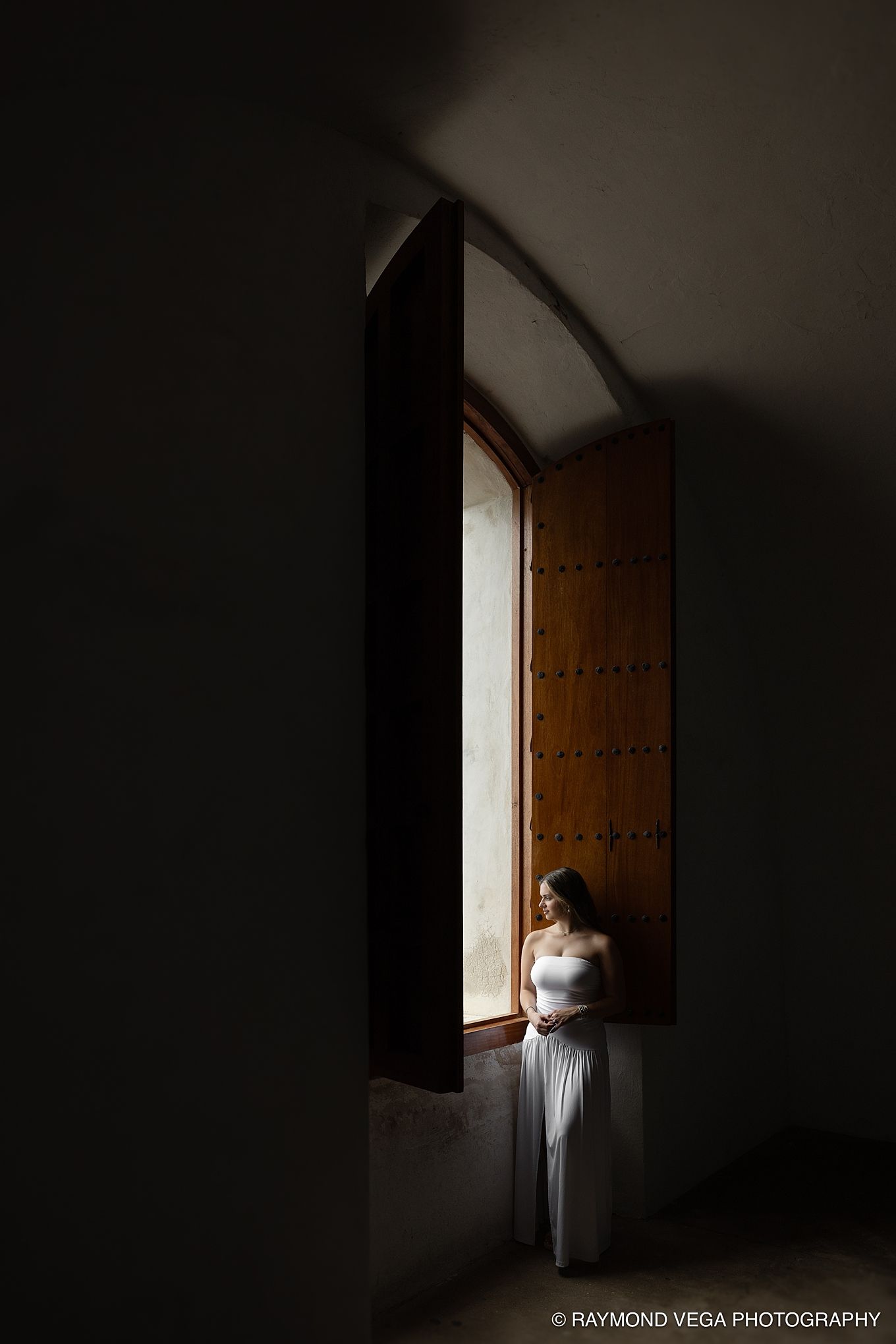 Bride in white gown standing by an arched wooden door in dramatic light, captured by Raymond Vega Photography.