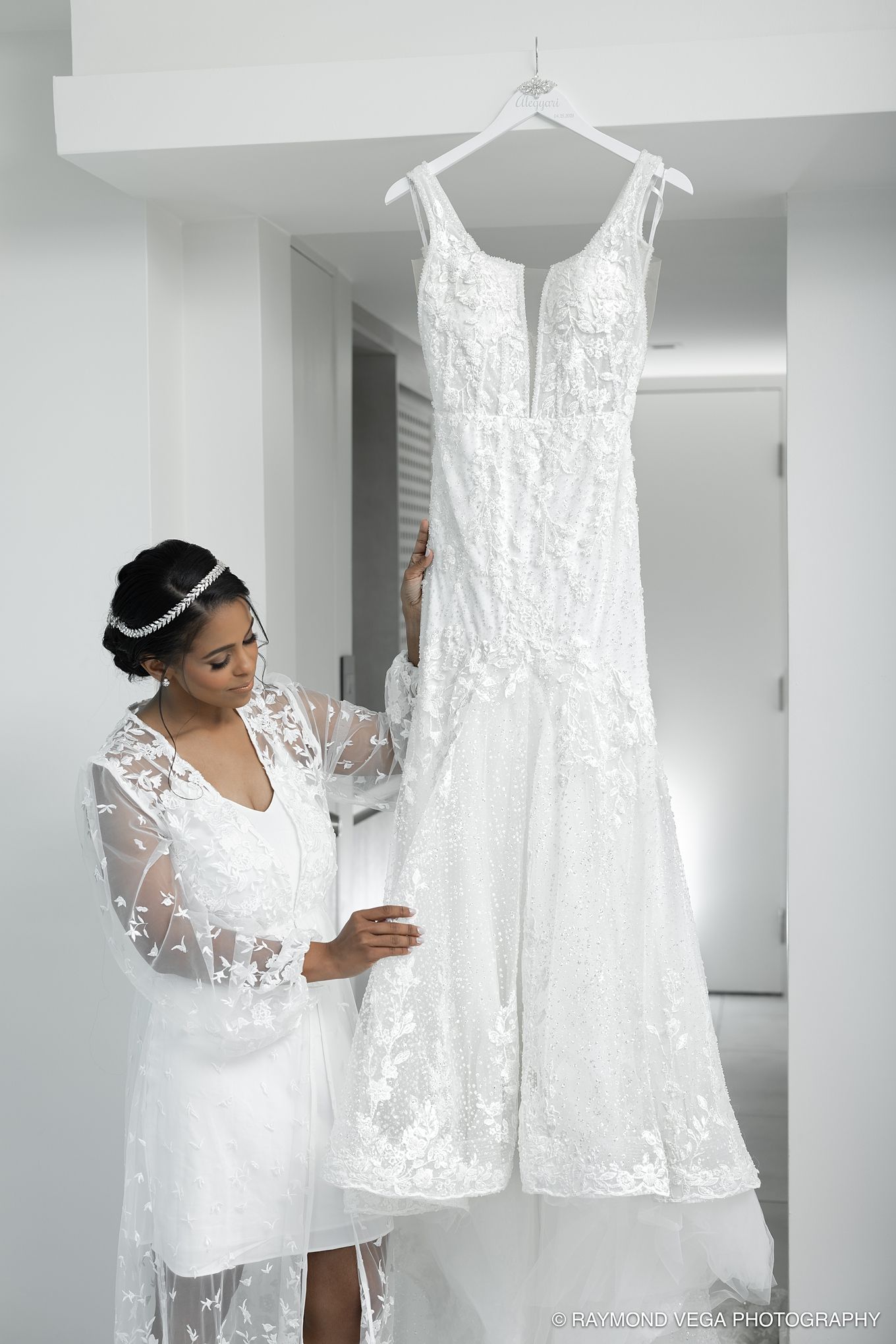 Bride in lace robe admiring her elegant white wedding gown hanging on a hanger in a bright white room.
