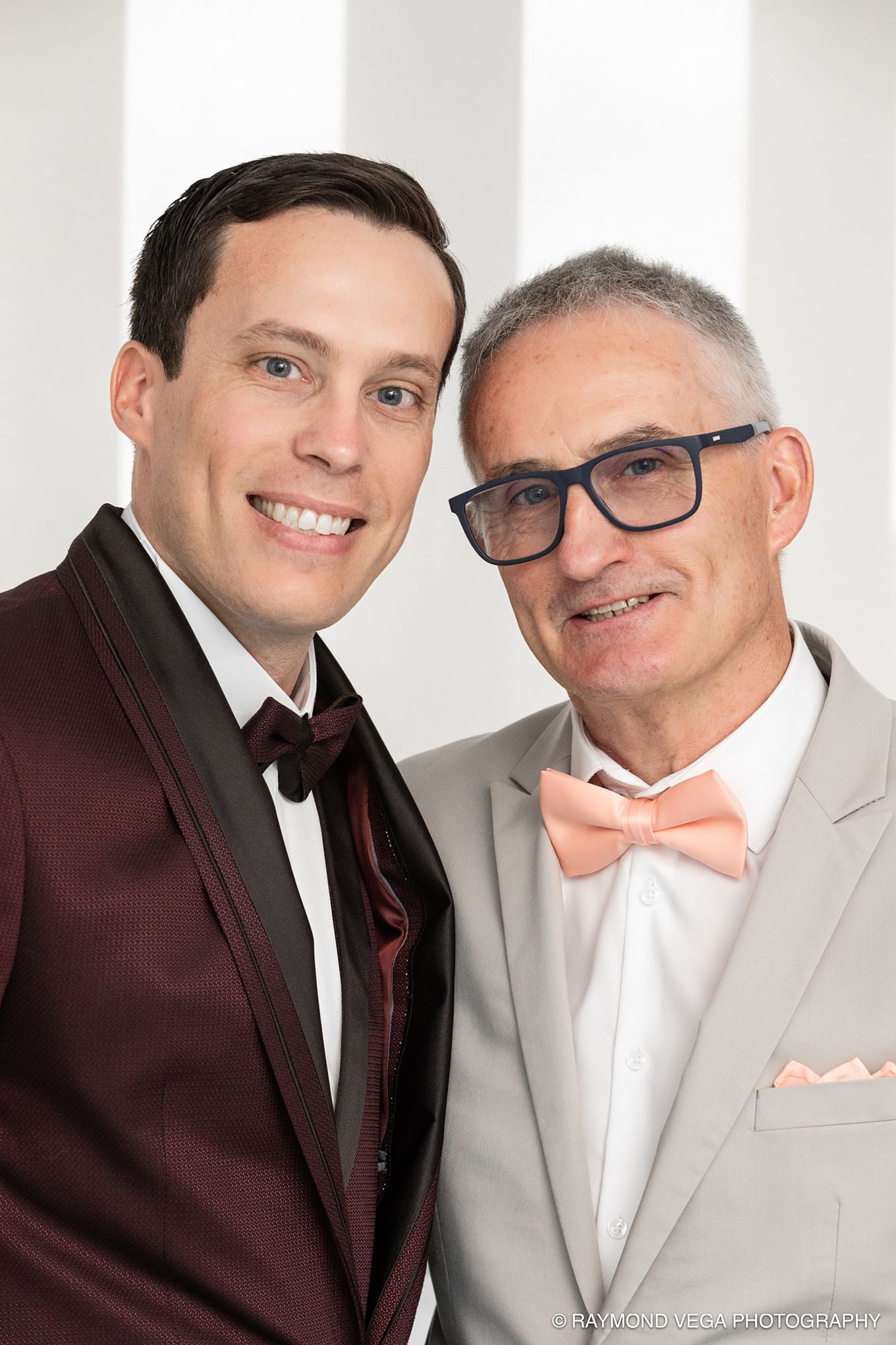 Two men in formal suits smiling together, one in burgundy tuxedo, one in gray suit with pink bow tie.