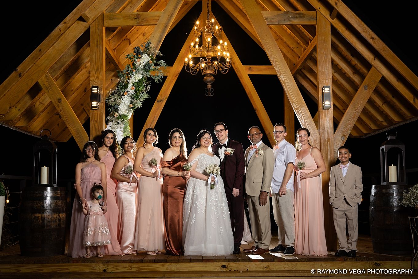 Wedding party poses under a wooden gazebo with chandelier at night, bride in white gown surrounded by bridesmaids in pink.