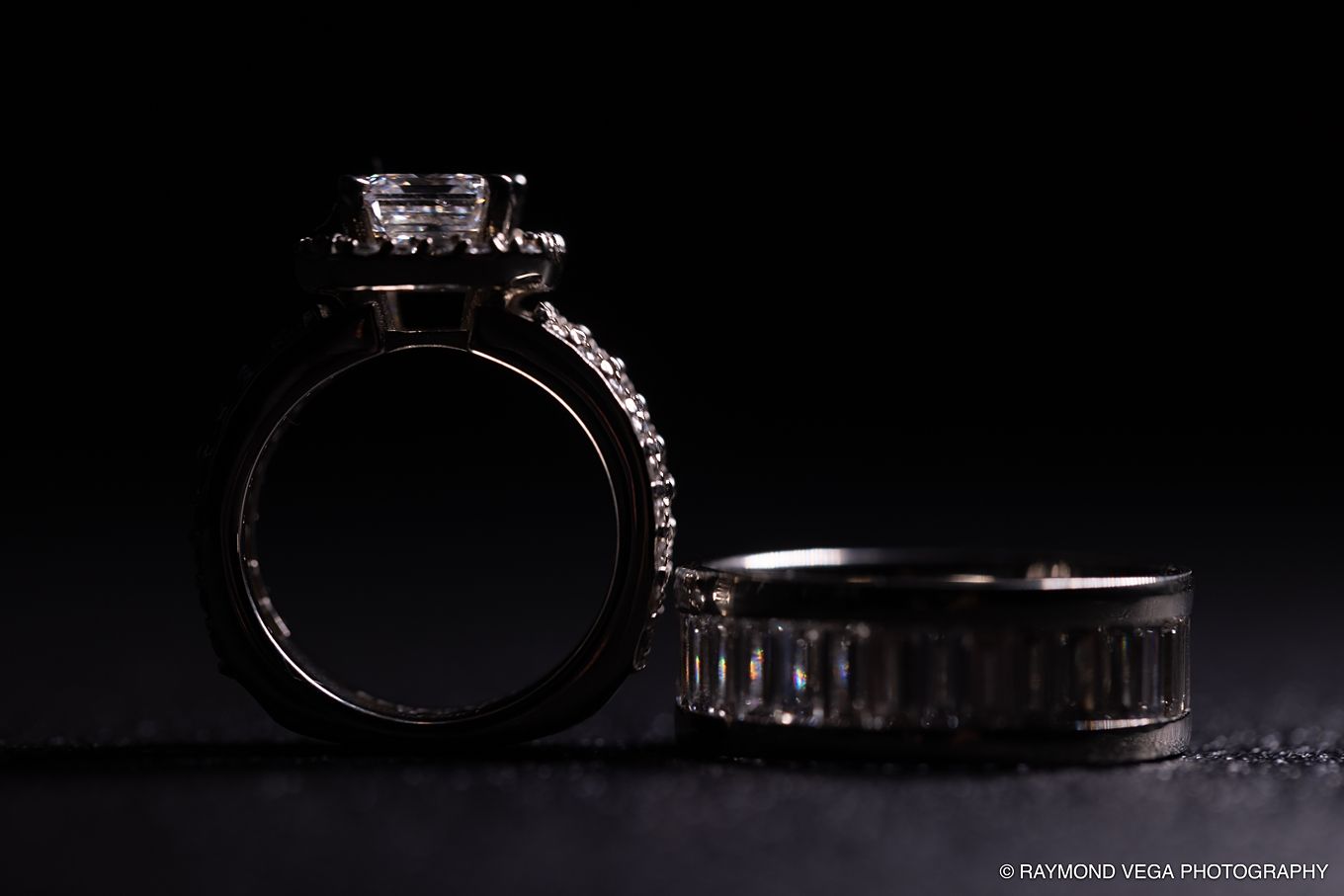 Two elegant diamond wedding rings, an engagement ring with halo setting and a band, photographed dramatically on dark background.