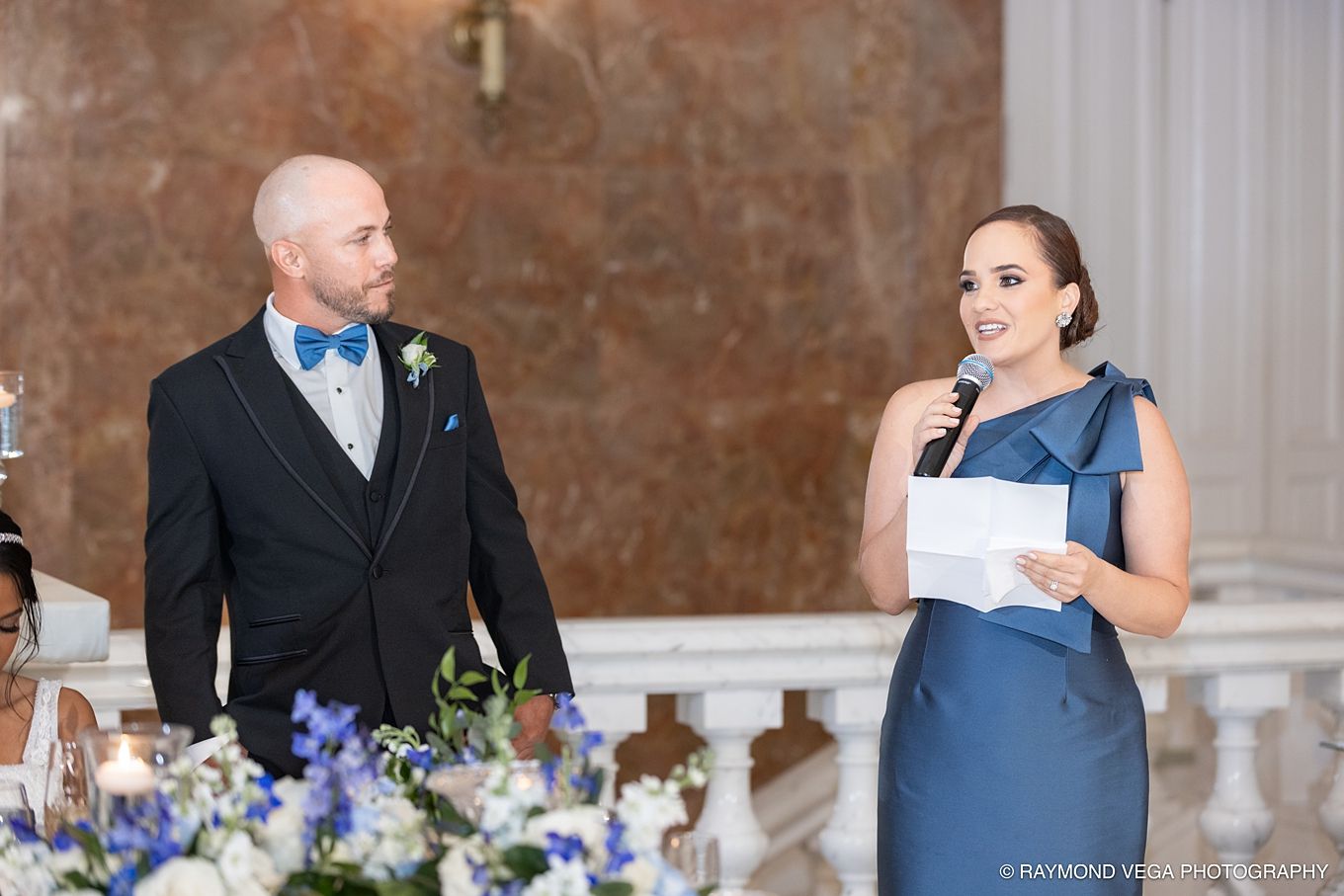 Bride in blue gown gives wedding speech beside groom in tuxedo at elegant reception venue.