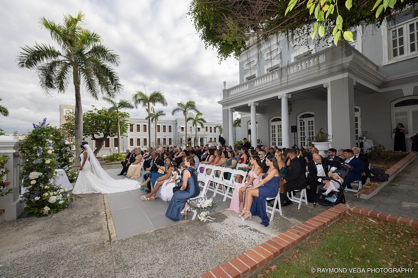 Outdoor wedding ceremony at elegant white colonial mansion with guests seated on white chairs surrounded by tropical palm trees.