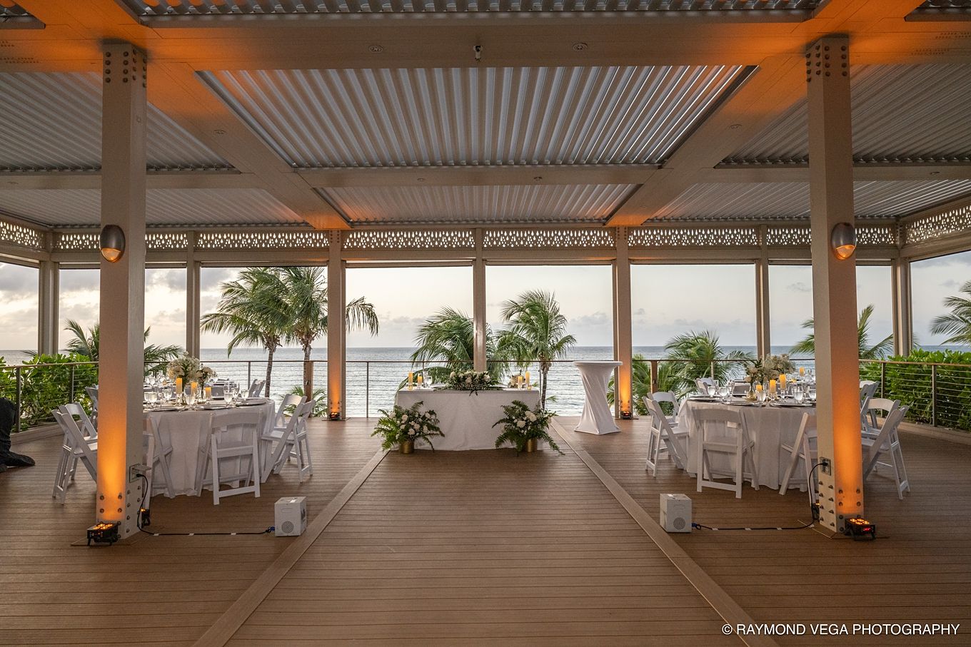Elegant outdoor wedding reception venue with white tables and chairs under a pergola overlooking a tropical ocean view. Courtyard by Marriot