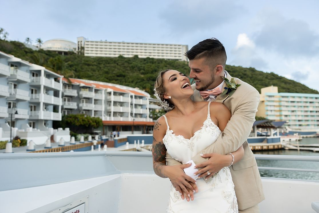 Bride and groom embracing on a boat with a tropical resort and lush green hillside in the background. El Conquistador Resort Wedding