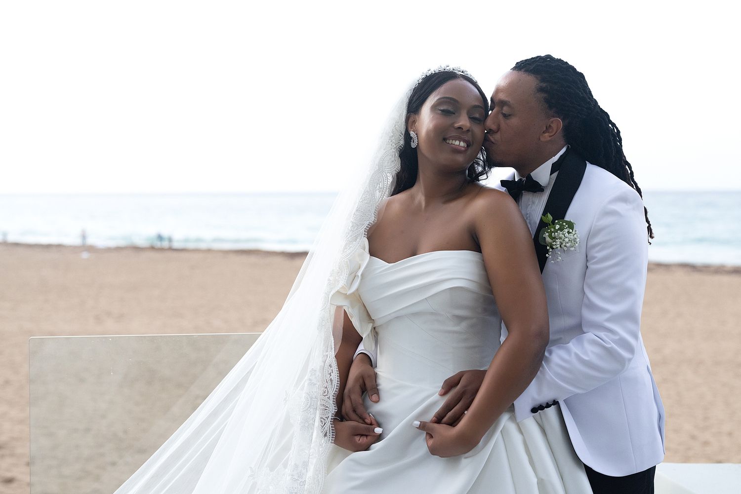 Couple in wedding attire on beach, bride in white gown with veil, groom in white tuxedo kissing her cheek. Hotel La Concha Puerto Rico