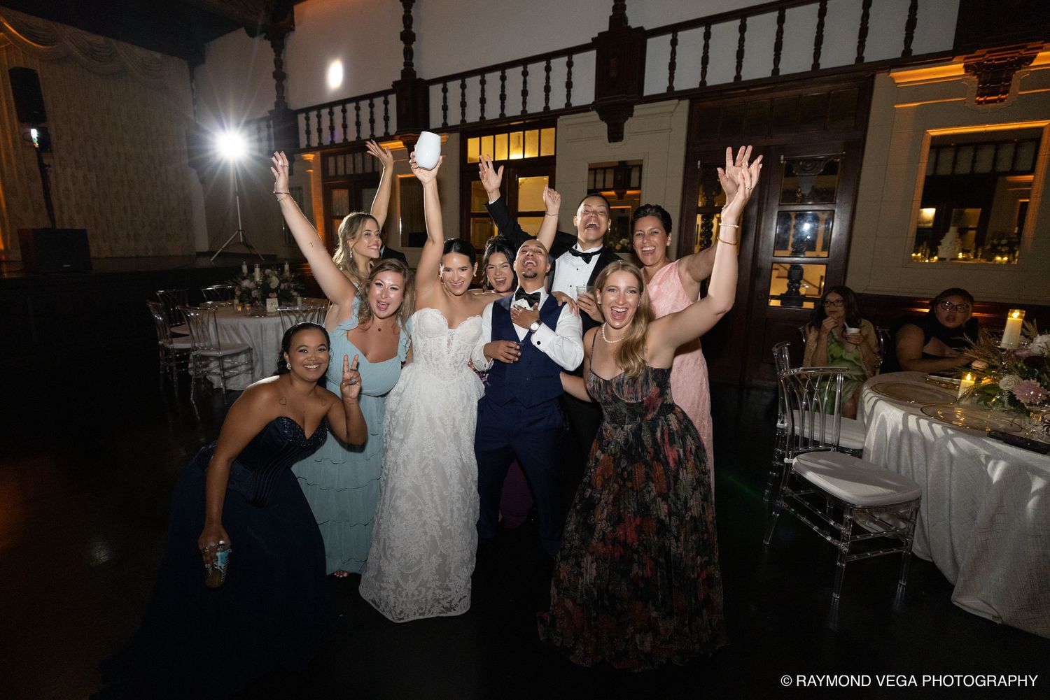 Joyful wedding party celebrating with raised hands and drinks at an elegant indoor reception venue.