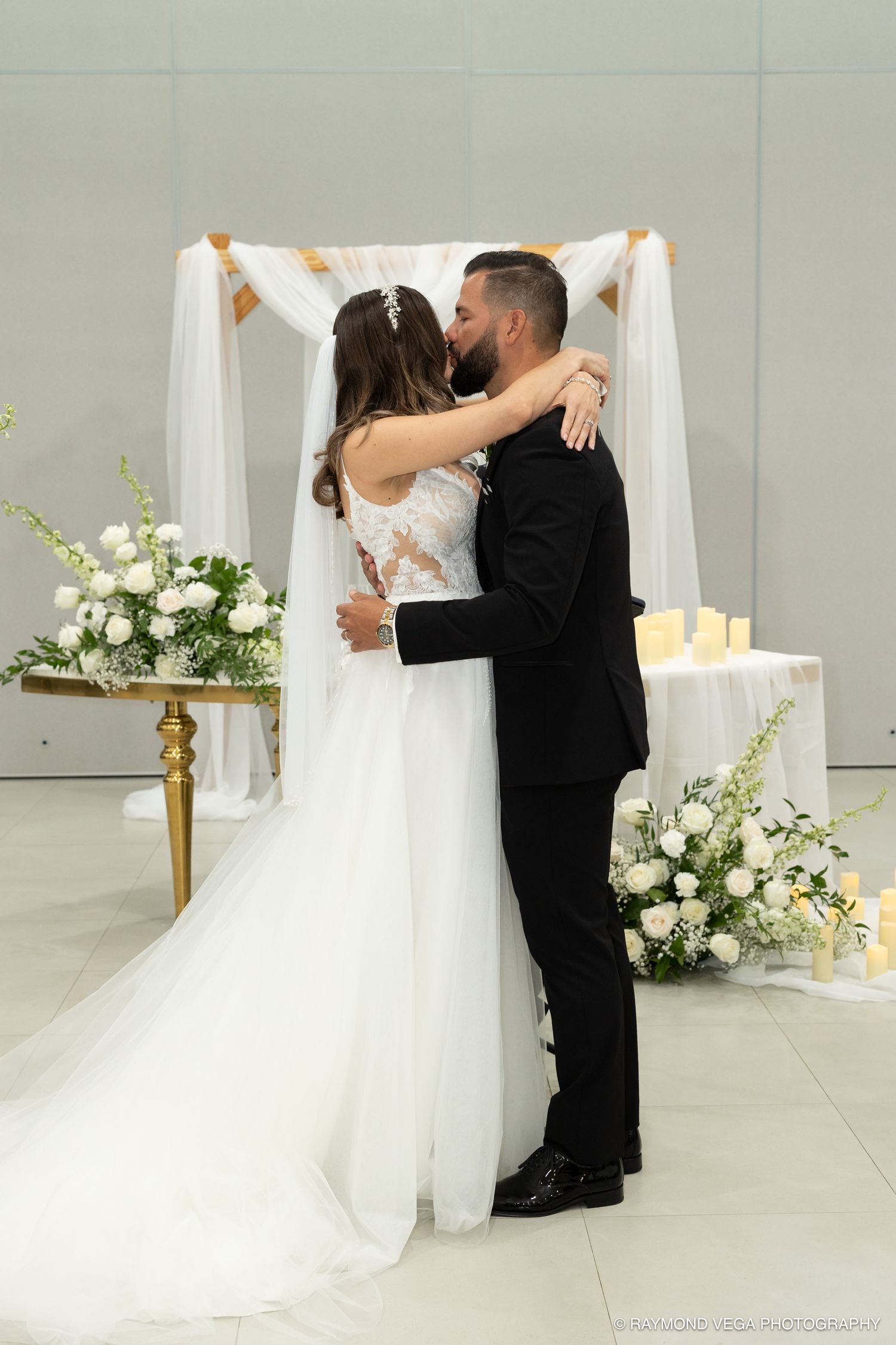 Bride and groom share their first kiss at the altar during an elegant wedding ceremony with white floral arrangements.