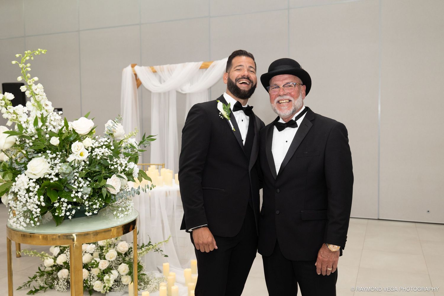 Two men in black tuxedos smiling at a wedding ceremony with white floral arrangements in the background.