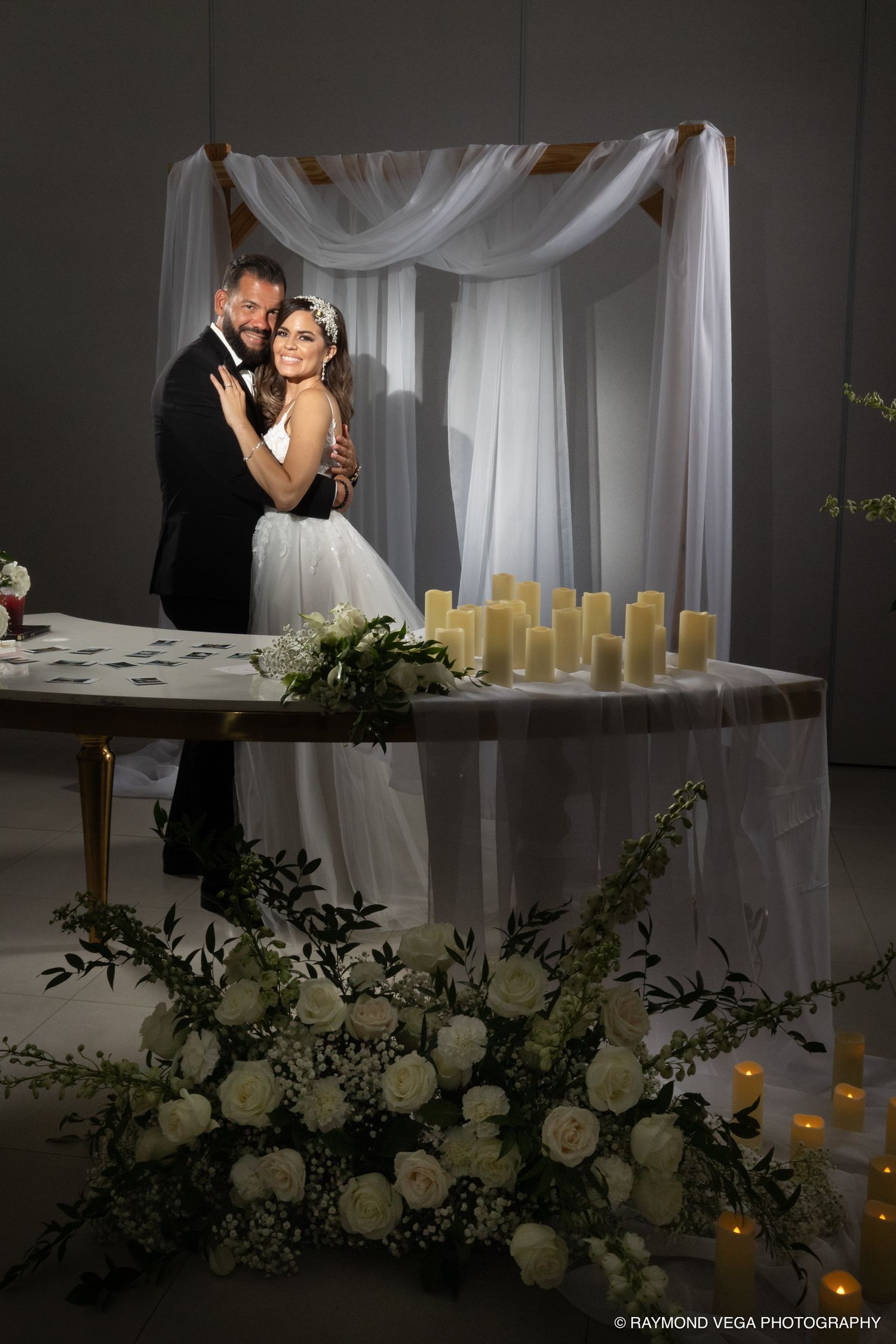 Bride and groom embrace before a draped white canopy altar with candles and white floral arrangements at elegant wedding ceremony.