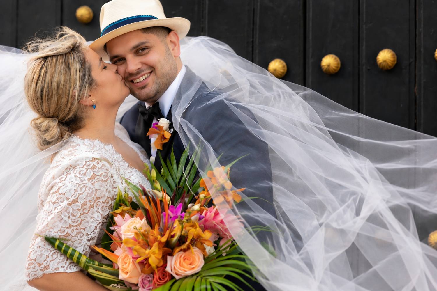 Bride and groom share a romantic moment, with veil flowing, as she kisses his cheek holding a tropical bouquet.