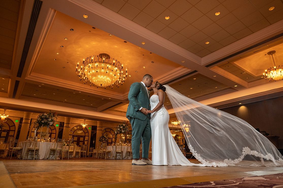 Bride and groom share romantic kiss on dance floor in elegant ballroom with chandeliers and draped wedding veil.