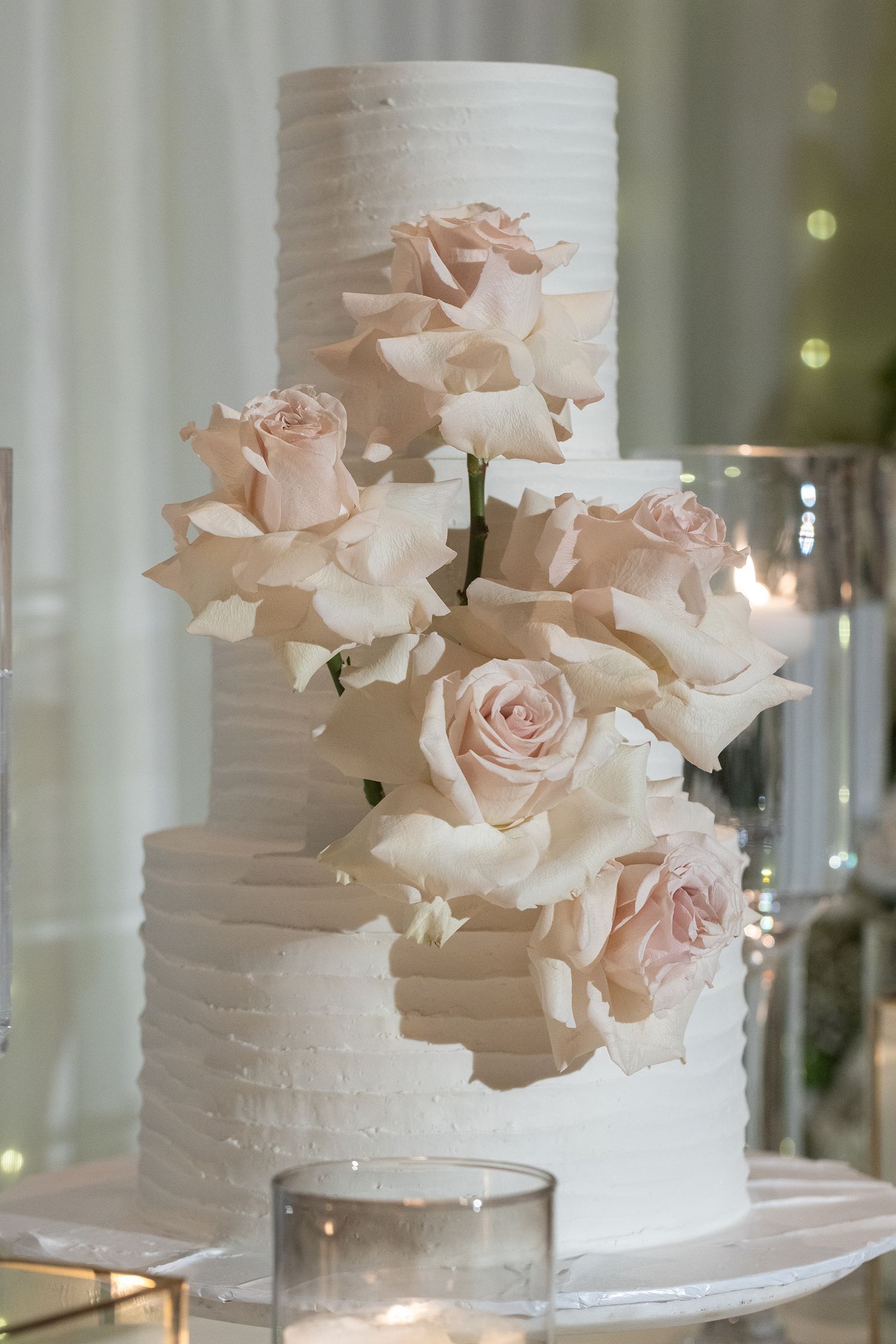 Elegant three-tier white wedding cake adorned with blush pink roses on a mirrored stand with candles.