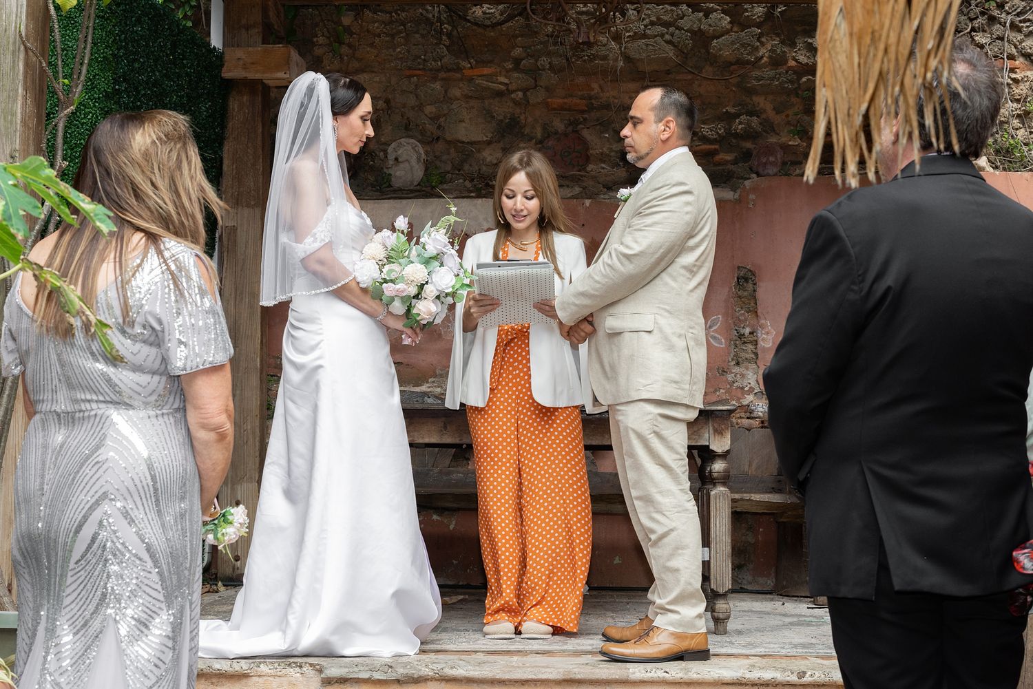 Bride and groom exchange vows outdoors with officiant in orange pants holding a book during an intimate wedding ceremony. AMR Destination