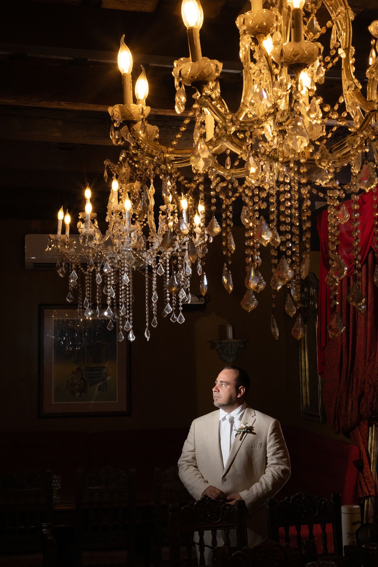 Groom in tan suit stands beneath ornate crystal chandelier in dimly lit elegant venue.