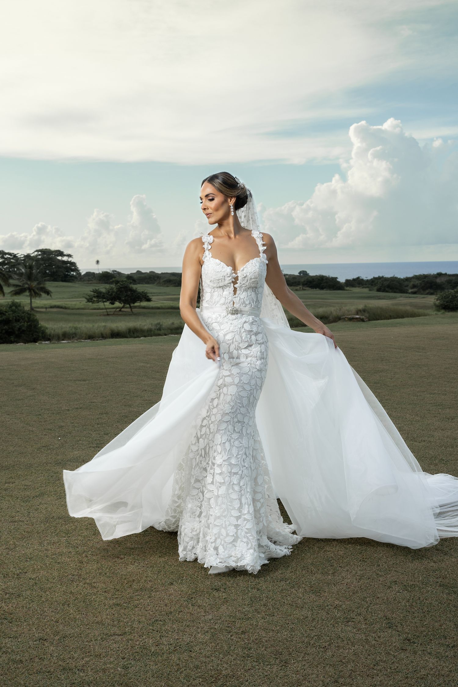 Elegant bride in a floral applique mermaid wedding gown with flowing veil poses outdoors against a cloudy sky.