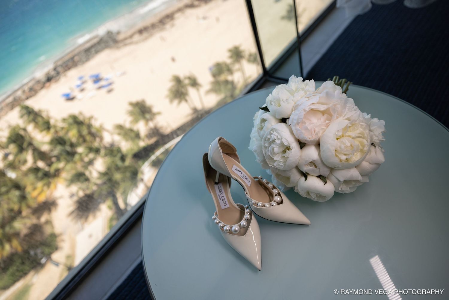 Elegant bridal heels and white peony bouquet on a table with a stunning beach view from a high-rise venue.