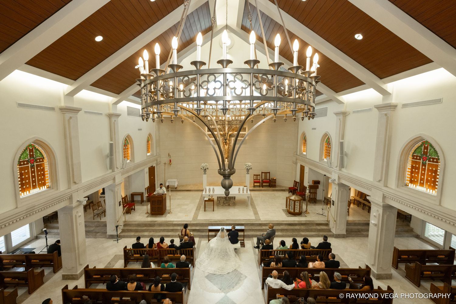 Elegant church wedding ceremony with ornate chandelier, stained glass windows, and bride in white gown walking down the aisle.