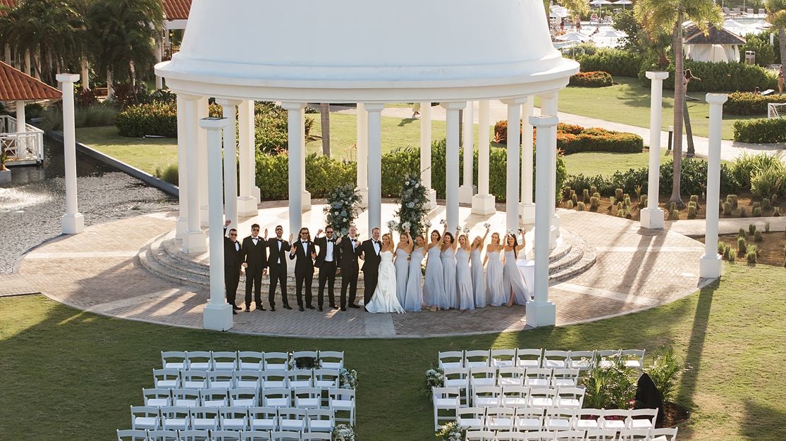 Wedding party poses under white gazebo at elegant outdoor ceremony venue with rows of chairs.